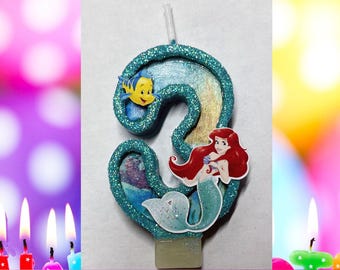 May include: A number 3 shaped birthday candle with a teal glitter border, featuring a small yellow fish cutout and a red-haired mermaid cutout. The text CUSTOM BIRTHDAY CANDLES and SUZDESIGNZ appears at the top and bottom of the image frame.