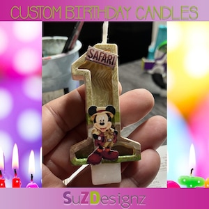 May include: A brown and tan birthday candle shaped like the number one with a safari theme. The candle has a picture of Mickey Mouse dressed in a safari outfit. The candle also has the word "Safari" written on it.