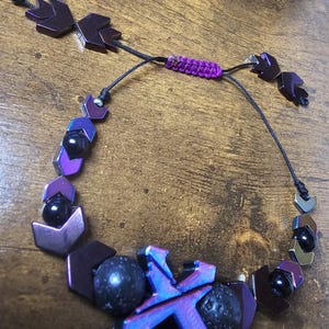 May include: A beaded bracelet featuring a mix of dark purple, black, and iridescent blue beads. The bracelet includes geometric shapes, round beads, and a central X-shaped design. The adjustable closure is a woven purple cord.