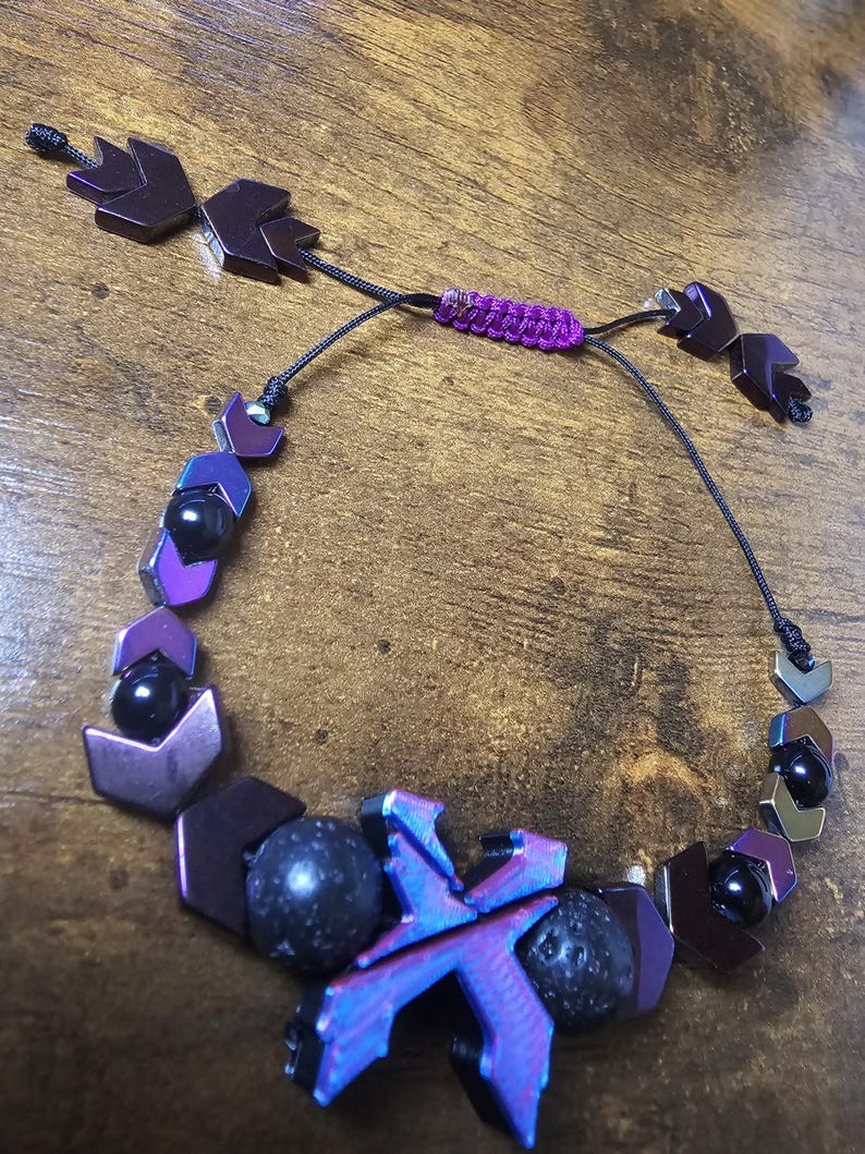 May include: A beaded bracelet featuring a mix of dark purple, black, and iridescent blue beads. The bracelet includes geometric shapes, round beads, and a central X-shaped design. The adjustable closure is a woven purple cord.