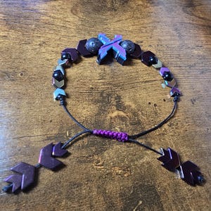 May include: A handmade bracelet featuring a mix of dark purple, black, and iridescent beads. The bracelet has a sliding knot closure with arrow-shaped end pieces. The central design includes a cross-shaped element with a mix of purple and blue hues.