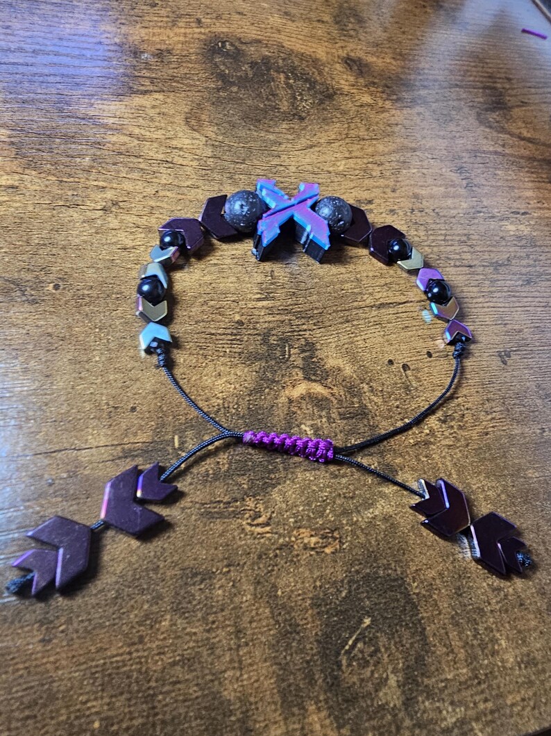 May include: A handmade bracelet featuring a mix of dark purple, black, and iridescent beads. The bracelet has a sliding knot closure with arrow-shaped end pieces. The central design includes a cross-shaped element with a mix of purple and blue hues.