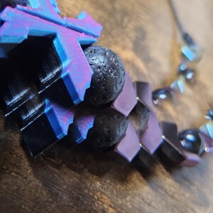May include: A close-up of a necklace with a geometric design. The necklace features black lava rock beads and purple, black, and blue geometric shapes. The necklace is on a wooden surface.