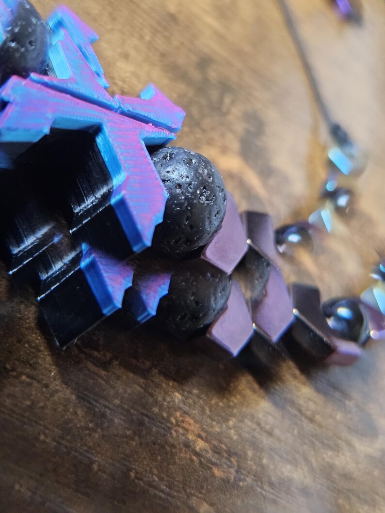May include: A close-up of a necklace with a geometric design. The necklace features black lava rock beads and purple, black, and blue geometric shapes. The necklace is on a wooden surface.