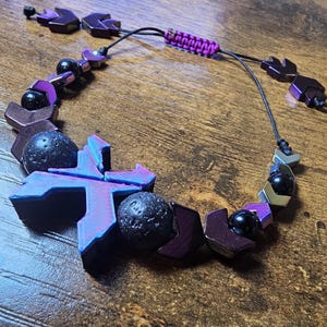 May include: A handmade bracelet featuring geometric purple and black beads, lava rock spheres, and a woven purple cord. The adjustable bracelet has a unique design with a mix of textures and colors.