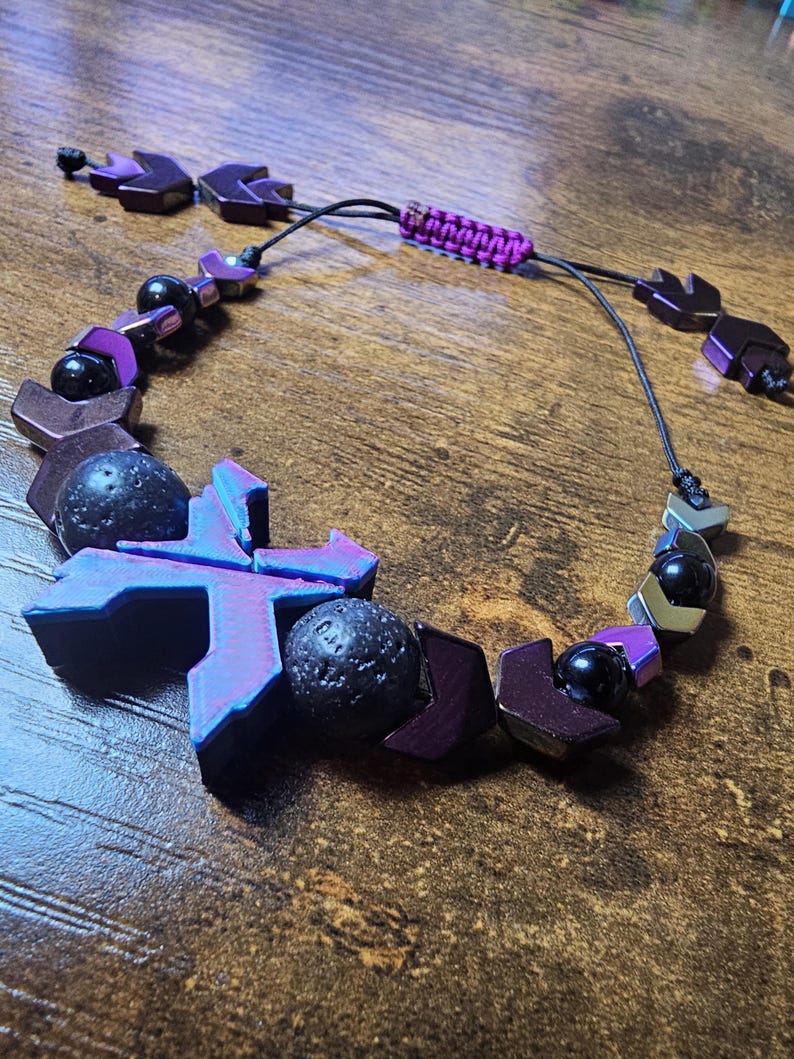 May include: A handmade bracelet featuring geometric purple and black beads, lava rock spheres, and a woven purple cord. The adjustable bracelet has a unique design with a mix of textures and colors.