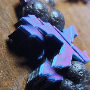 May include: Close-up of a blue and purple X-shaped object with a layered design, resting on a wooden surface. The object is paired with several dark, spherical, textured stones.