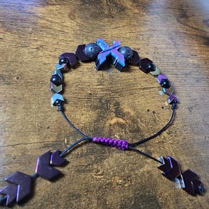 May include: Adjustable bracelet featuring a central iridescent X-shaped bead, flanked by dark purple and black beads. The bracelet includes black cord and purple accents, with leaf-shaped end pieces. A unique, handcrafted jewelry item.