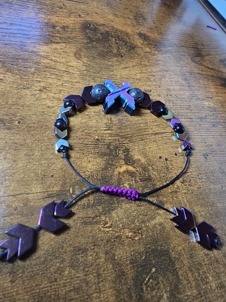 May include: Adjustable bracelet featuring a central iridescent X-shaped bead, flanked by dark purple and black beads. The bracelet includes black cord and purple accents, with leaf-shaped end pieces. A unique, handcrafted jewelry item.