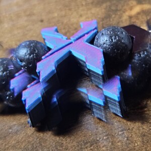 May include: A close-up of a bracelet featuring a layered, multi-colored letter "X" pendant with blue, purple, and black hues. The bracelet also includes several dark, textured beads. The background is a blurred, brown surface.