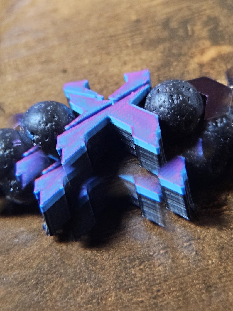 May include: A close-up of a bracelet featuring a layered, multi-colored letter "X" pendant with blue, purple, and black hues. The bracelet also includes several dark, textured beads. The background is a blurred, brown surface.