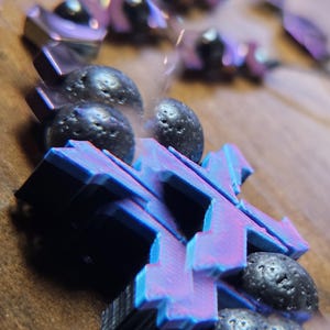 May include: Close-up of a bracelet featuring geometric, blue and purple shapes and dark, textured beads. The bracelet is set against a blurred, neutral background, highlighting the contrast of colors and textures.