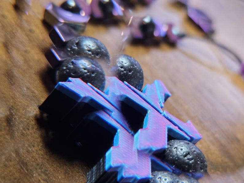 May include: Close-up of a bracelet featuring geometric, blue and purple shapes and dark, textured beads. The bracelet is set against a blurred, neutral background, highlighting the contrast of colors and textures.