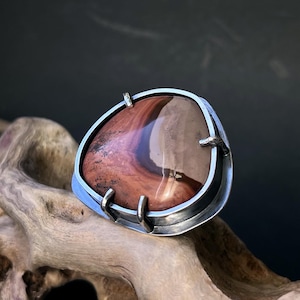 Könnte beinhalten: Ein silberner Ring mit einem großen, unregelmäßig geformten, rot-braunen Achat-Stein, der in der Mitte eingesetzt ist. Der Ring liegt auf einem Stück Treibholz.