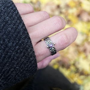 Puede incluir: Anillo de plata martillada con una pequeña gema redonda y morada. El anillo tiene una superficie texturizada y se lleva en un dedo. El fondo está borroso, con toques de amarillo y verde.