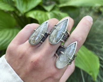 May include: Three silver rings with large, teardrop-shaped stones. The stones have a mix of white, gray, and brown patterns. The rings have a textured band and gold accents, creating a unique and artistic look.