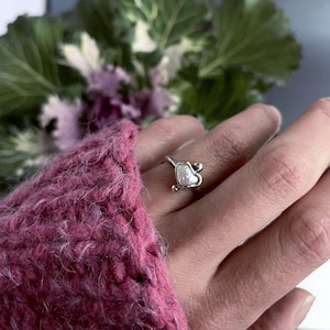 May include: A silver ring with a heart-shaped pearl. The ring is on a finger and the background is blurred.