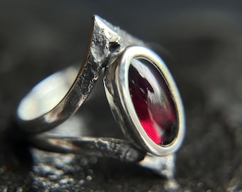 May include: A silver ring with a textured band and a red gemstone set in a bezel setting.