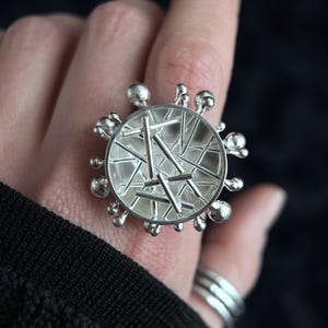 May include: A silver ring with a circular design. The ring has a central geometric pattern of intersecting lines and is surrounded by small, spherical accents. The ring is worn on a finger, with a dark background.