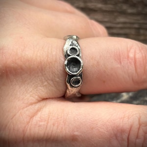 May include: A silver ring with a textured, organic design. The ring features three circular, recessed details. The metal has a rough, uneven surface, giving it a handcrafted appearance. The ring is worn on a finger.