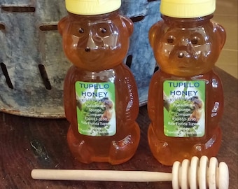 May include: Two clear glass bear-shaped bottles filled with honey. The bottles have yellow lids and labels that say "Tupelo Honey" and include the company name, phone number, and website. A wooden honey dipper is in the foreground.