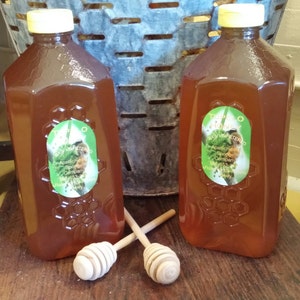 May include: Two clear glass bottles filled with honey, labeled "Tupelo Raw Unfiltered Honey" with a bee and honeycomb design. The bottles are sitting on a wooden surface with two wooden honey dippers.