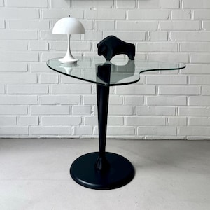 Peut inclure: Une table d'appoint moderne avec un plateau en verre transparent et un pied noir. Une sculpture de taureau noir et une lampe blanche sont posées sur la table. Une estampe en noir et blanc encadrée est accrochée au mur de briques blanches.