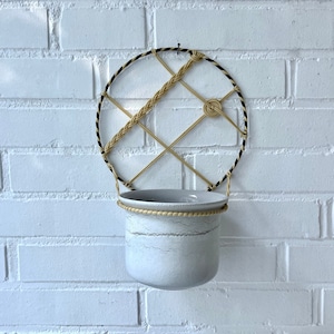 May include: A white ceramic planter with a rope trim hangs from a black and beige rope macrame wall hanging.