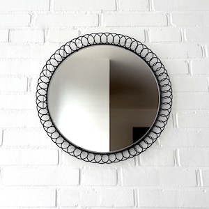 Può includere: Uno specchio rotondo con una cornice in metallo nero con un motivo ovale ripetuto. Lo specchio è montato su un muro di mattoni bianchi, riflettendo lo spazio circostante. La cornice aggiunge un tocco decorativo.