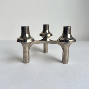 May include: A silver-colored metal candle holder with three candle cups. The candle holder has a unique design with a curved base and three legs. The cups are wide at the top and narrow at the base.