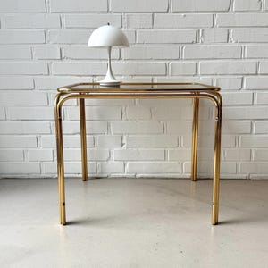 May include: A gold-toned side table with a clear glass top and a white mushroom-shaped lamp. The table has a rectangular shape with rounded corners and straight legs. The lamp is centered on the table.