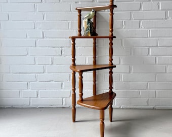 May include: A tall, four-tiered corner shelf unit crafted from medium-toned wood with turned spindle supports. The shelves are triangular in shape, and a small, decorative ceramic bird figurine is placed on the second shelf from the top.