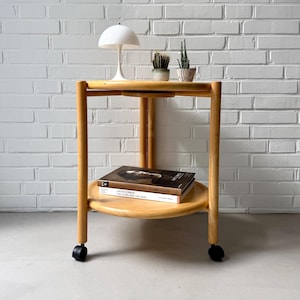 Può includere: Un tavolino a due livelli in legno chiaro, con ripiano superiore e inferiore rotondi. Il tavolo ha una lampada bianca, due piccole piante in vaso e due libri. Il tavolo è su ruote ed è appoggiato a un muro di mattoni bianchi.