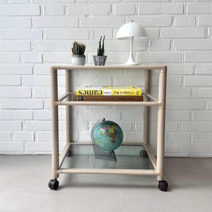 May include: A beige rolling side table with glass shelves. The top shelf holds two small potted plants and a white lamp. A yellow book and a globe are on the lower shelves. The table is against a white brick wall.