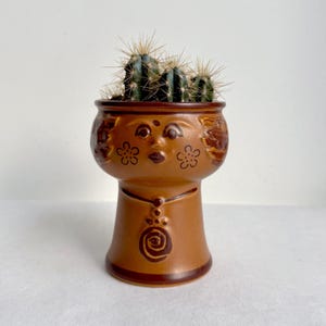 Könnte beinhalten: Ein brauner Keramik-Blumentopf mit Gesichtsdesign, der einen kleinen Kaktus enthält. Der Topf hat ein detailliertes Gesicht mit floralen und geometrischen Mustern. Der Kaktus hat grüne Stiele und scharfe Stacheln. Der Topf ist etwa 15 cm hoch.