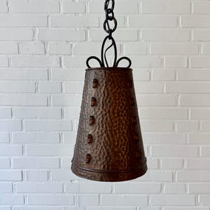 Könnte beinhalten: Eine hängende, glockenförmige Pendelleuchte mit gehämmertem Kupferfinish. Die Leuchte wird von einer schwarzen Kette und dekorativen Metallarbeiten abgehängt. Der Kupferschirm hat eine strukturierte Oberfläche und ist mit Nieten verziert.