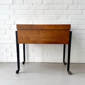 May include: A mid-century modern wooden cabinet with a rectangular shape. The cabinet is made of light brown wood and has a black metal frame with four small wheels. The cabinet is set against a white brick wall.