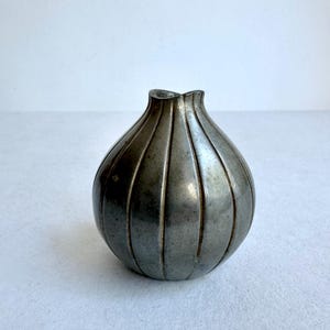 May include: A small, antique-style vase with a bulbous shape and a narrow, fluted neck. The vase is made of a dark, metallic material with vertical grooves running from the base to the top. The overall design is simple and elegant, suitable for a minimalist aesthetic.