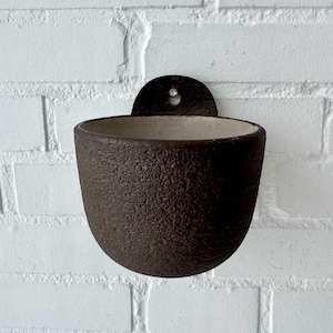 Könnte beinhalten: Ein dunkelbrauner, strukturierter Wandtopf aus Keramik mit abgerundeter Form. Der Topf hat eine kleine, kreisförmige Montageplatte mit einem Loch zum Aufhängen. Der Innenraum ist heller und neutral.