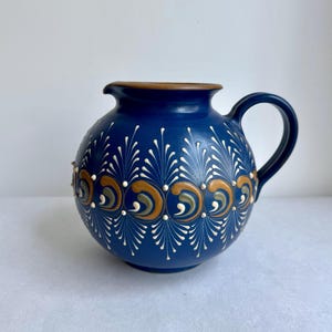 Op de afbeelding: Een ronde, donkerblauwe keramische kan met een handvat en tuit. De kan is versierd met witte, gouden en groene bloempatronen. De rand is bruin.