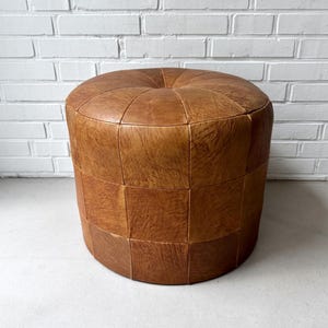 May include: A round, tan-coloured leather ottoman featuring a tufted top centre and a segmented, panelled construction. The surface displays a natural, textured grain pattern across the cylindrical body, creating a uniform and structured appearance.