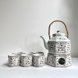 May include: A white ceramic tea set with a brown floral pattern. The set includes a teapot with a woven handle, a warmer, and six matching cups. The teapot and cups have a decorative brown border.