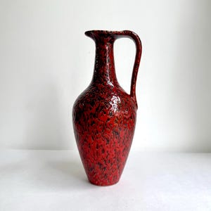 May include: A tall ceramic pitcher featuring a vibrant red glaze with a textured, mottled black pattern throughout. The vessel has a slender neck, a flared rim, and a single curved handle extending from the neck to the upper body of the piece.