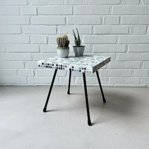 May include: A square mosaic tile side table with a black metal frame. The tabletop features a pattern of small, square tiles in shades of white, gray, and teal. Two potted plants sit on top of the table. The table is approximately 18 inches wide.