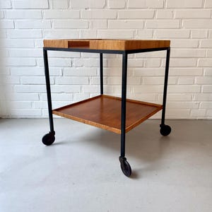 May include: A two-tiered rolling cart featuring a black metal frame and four casters. The top tier has a light brown textured rattan-wrapped rim, while the bottom tier consists of a flat, smooth, medium-brown wooden shelf surface.