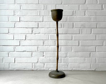 May include: A tall, brass plant stand with a bamboo-style stem and a round base. The stand is in front of a white brick wall.