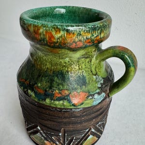 K&ouml;nnte beinhalten: Eine Keramiktasse mit gr&uuml;nem Innenraum und Henkel. Die Au&szlig;enseite weist ein strukturiertes Design mit Gr&uuml;nt&ouml;nen, Orange und Braun auf. Der Boden hat ein geschnitztes geometrisches Muster. Die Tasse ist etwa 13 cm hoch.