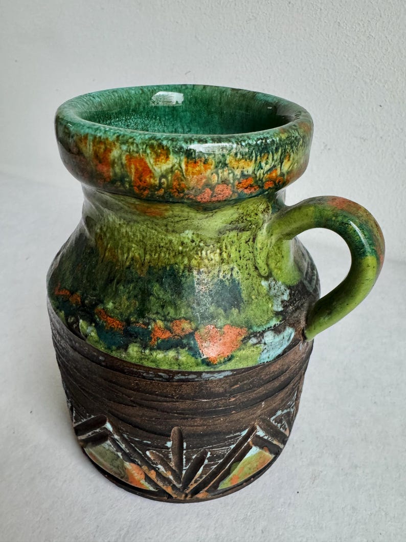 K&ouml;nnte beinhalten: Eine Keramiktasse mit gr&uuml;nem Innenraum und Henkel. Die Au&szlig;enseite weist ein strukturiertes Design mit Gr&uuml;nt&ouml;nen, Orange und Braun auf. Der Boden hat ein geschnitztes geometrisches Muster. Die Tasse ist etwa 13 cm hoch.