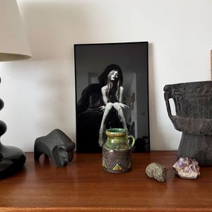 K&ouml;nnte beinhalten: Ein Stillleben-Arrangement mit einem gerahmten Schwarz-Wei&szlig;-Foto einer Frau, einer kleinen Keramikvase mit gr&uuml;n-brauner Glasur, einer schwarzen Tierskulptur und dekorativen Steinen auf einer Holzoberfl&auml;che.