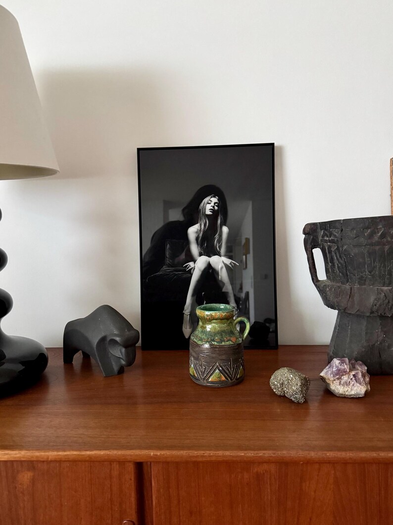 K&ouml;nnte beinhalten: Ein Stillleben-Arrangement mit einem gerahmten Schwarz-Wei&szlig;-Foto einer Frau, einer kleinen Keramikvase mit gr&uuml;n-brauner Glasur, einer schwarzen Tierskulptur und dekorativen Steinen auf einer Holzoberfl&auml;che.